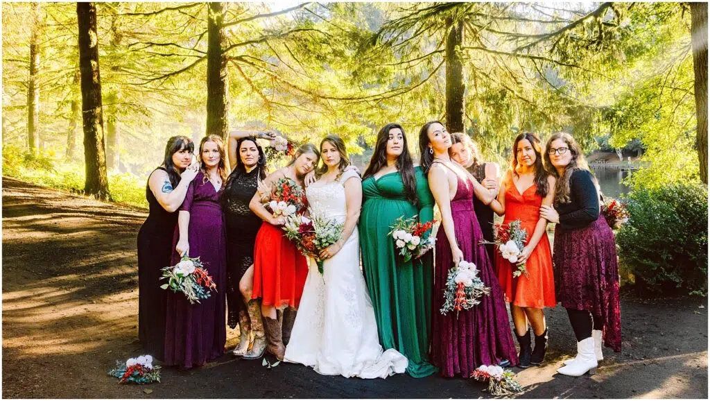 Snohomish Wedding Photographer GSquared Weddings A group of women, including a bride in a white gown, stand in a forest, surrounded by sunlight filtering through the trees. They wear colorful dresses in various shades, holding bouquets. The scene is warm and vibrant with lush greenery. Serving Seattle, Snohomish and Orlando