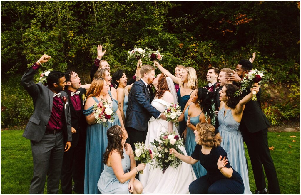 Snohomish Wedding Photographer GSquared Weddings A wedding party poses outdoors with the bride and groom kissing in the center. Bridesmaids in blue dresses and groomsmen in dark suits cheer and hold bouquets against a green, leafy background. Everyone appears joyful and celebratory. Serving Seattle, Snohomish and Orlando