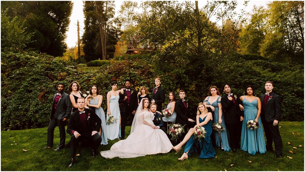 Snohomish Wedding Photographer GSquared Weddings A wedding party poses outdoors on a lawn against a backdrop of trees and greenery. The bride sits in the center in a white gown, surrounded by bridesmaids in blue dresses and groomsmen in dark suits. The atmosphere is elegant and relaxed. Serving Seattle, Snohomish and Orlando