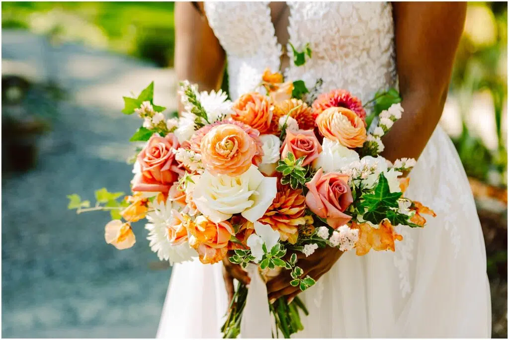 Snohomish Wedding Photographer GSquared Weddings A bride holds a vibrant bouquet of orange, pink, and white flowers mixed with greenery. She wears a white lace dress. The background is a blurred outdoor scene with green foliage. Serving Seattle, Snohomish and Orlando