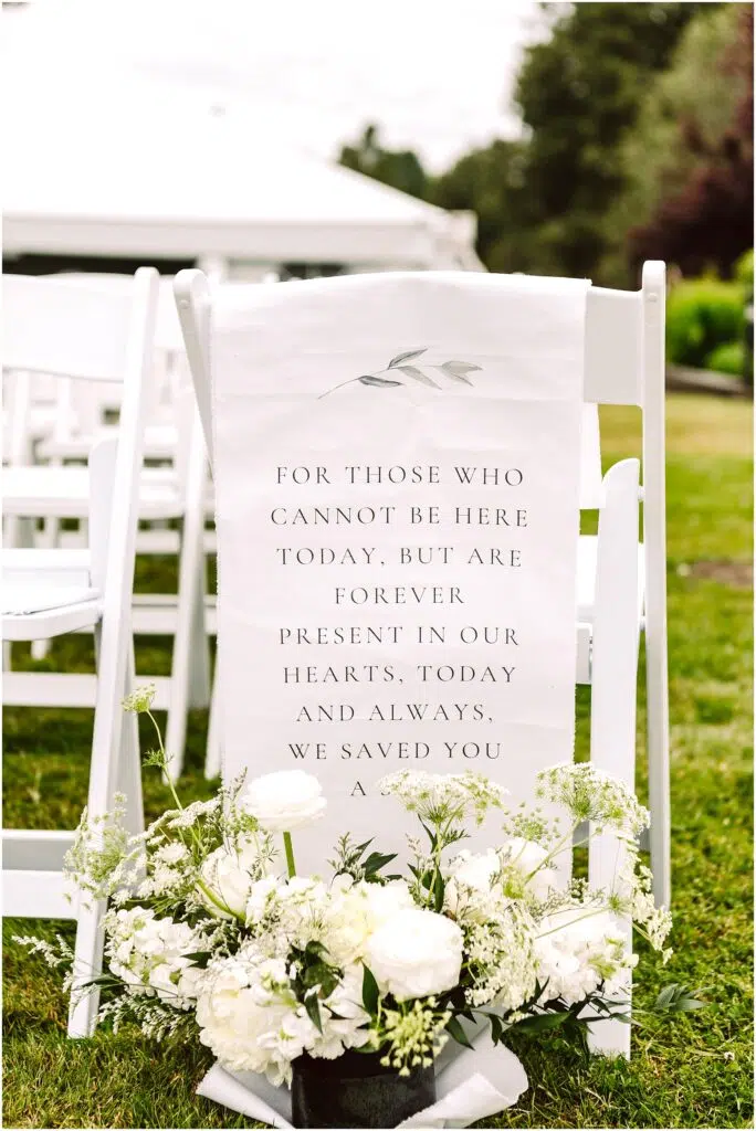Snohomish Wedding Photographer GSquared Weddings A wedding chair with a white cloth draped over the back, displaying a message memorializing absent loved ones. Below, a bouquet of white flowers and greenery is placed on the grass. White chairs and a tent are in the background. Serving Seattle, Snohomish and Orlando
