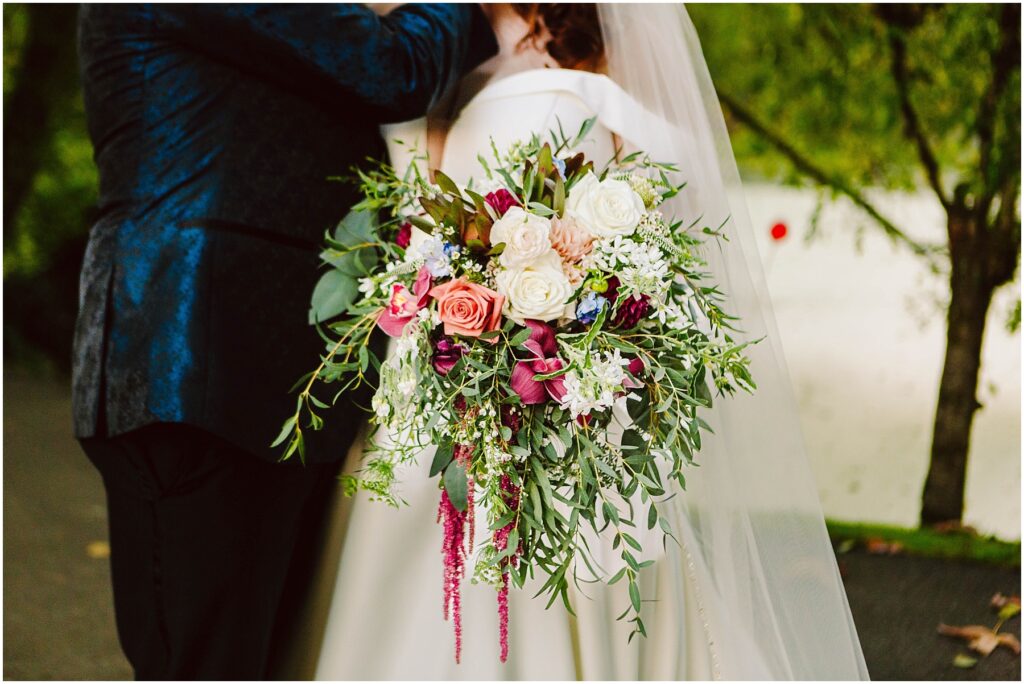 Snohomish Wedding Photographer GSquared Weddings Bride holding a large, vibrant bouquet of pink, white, and purple flowers with green foliage. She stands beside a person in a dark suit. Background shows trees and a white surface, possibly a building or wall. Serving Seattle, Snohomish and Orlando