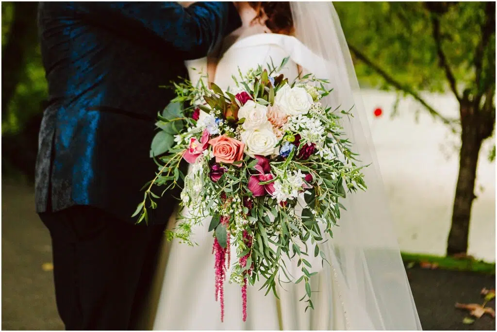 Snohomish Wedding Photographer GSquared Weddings Bride holding a large, vibrant bouquet of pink, white, and purple flowers with green foliage. She stands beside a person in a dark suit. Background shows trees and a white surface, possibly a building or wall. Serving Seattle, Snohomish and Orlando