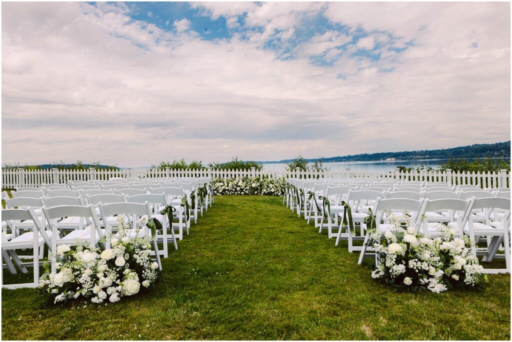 Snohomish Wedding Photographer GSquared Weddings A scenic outdoor wedding setup features two rows of white chairs lined with white floral arrangements on a grassy area. A white picket fence overlooks a body of water and a cloudy sky, providing a picturesque backdrop. Serving Seattle, Snohomish and Orlando