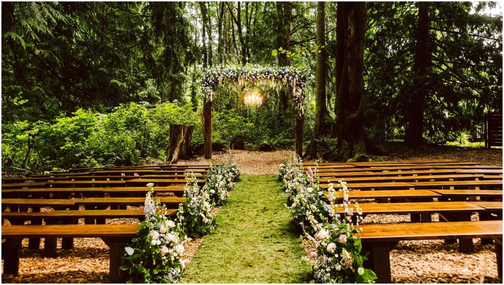 Snohomish Wedding Photographer GSquared Weddings A serene outdoor wedding setup in a forest. Wooden benches line a grassy aisle adorned with floral arrangements. An arch decorated with flowers stands at the end, surrounded by lush green trees and soft sunlight filtering through the leaves. Serving Seattle, Snohomish and Orlando