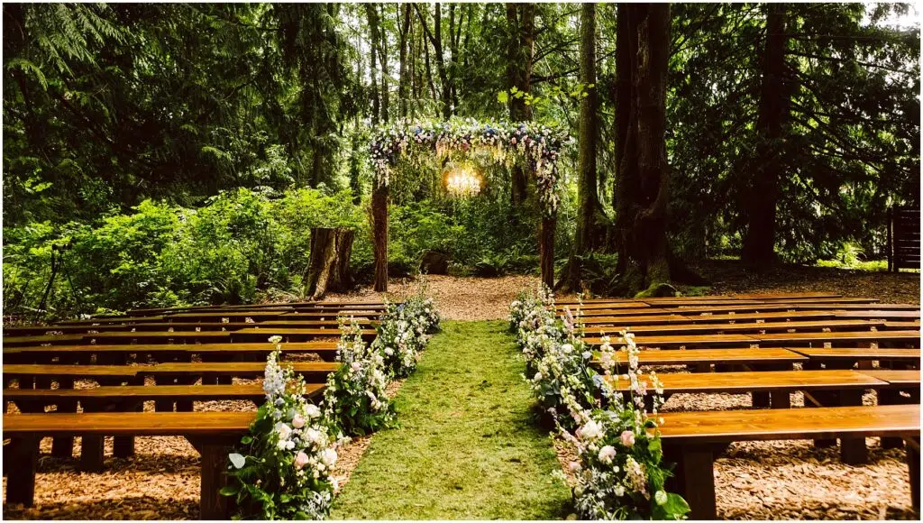 Snohomish Wedding Photographer GSquared Weddings A serene outdoor wedding setup in a forest. Wooden benches line a grassy aisle adorned with floral arrangements. An arch decorated with flowers stands at the end, surrounded by lush green trees and soft sunlight filtering through the leaves. Serving Seattle, Snohomish and Orlando