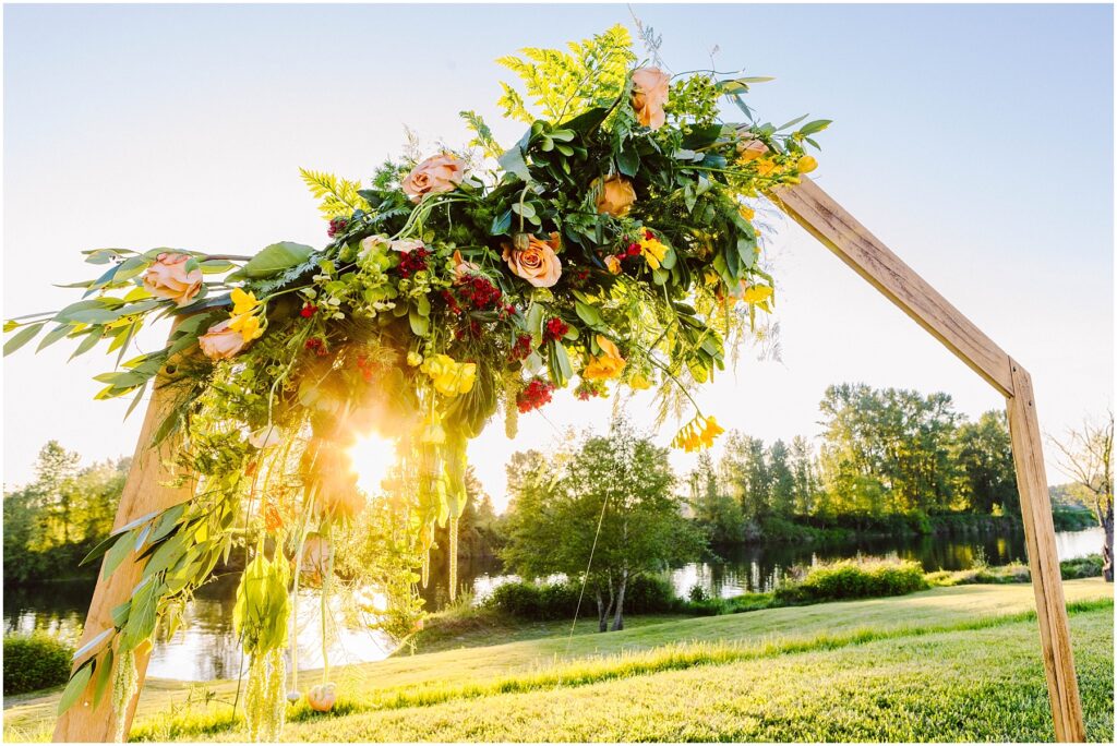 Snohomish Wedding Photographer GSquared Weddings A wooden arch adorned with green leaves, pink and yellow flowers, with sunlight shining through. The arch is set in a grassy area near a calm river, and trees are visible in the background under a bright blue sky. Serving Seattle, Snohomish and Orlando