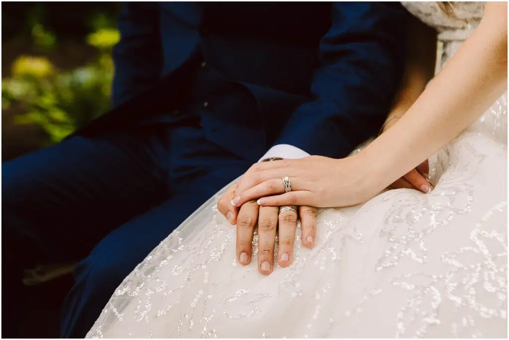 Snohomish Wedding Photographer GSquared Weddings Close-up of a couple sitting, holding hands. The person on the left is wearing a dark blue suit, and the person on the right is in a white, sequined dress with a wedding ring visible. The background is softly blurred with greenery. Serving Seattle, Snohomish and Orlando