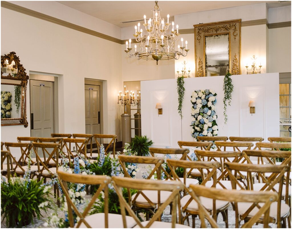 Snohomish Wedding Photographer GSquared Weddings Elegant wedding ceremony room with wooden chairs facing a white wall adorned with floral arrangements. Chandeliers and mirrors add sophistication, while greenery accents the space. Neutral tones create a serene atmosphere. Serving Seattle, Snohomish and Orlando