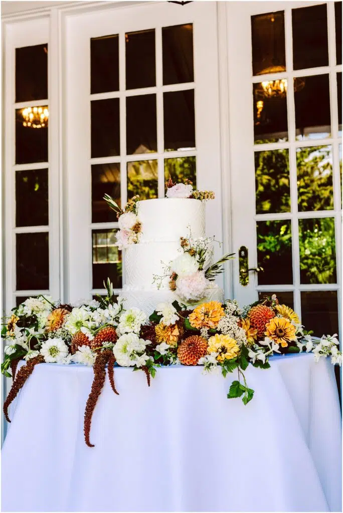 Snohomish Wedding Photographer GSquared Weddings A white, two-tier wedding cake adorned with pink and white flowers sits on a table covered with a white cloth. The table is decorated with a variety of colorful flowers and greenery, against a backdrop of white French doors with glass panes. Serving Seattle, Snohomish and Orlando