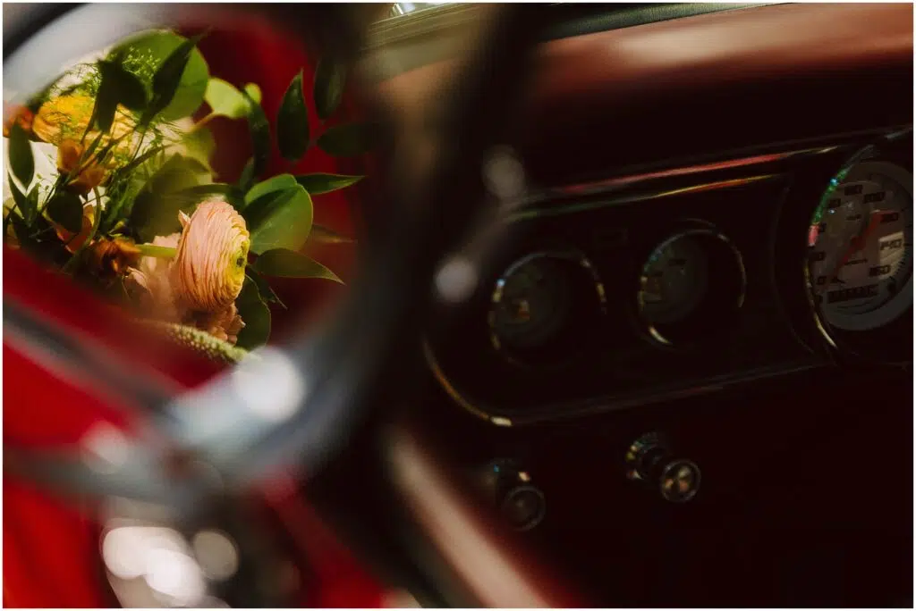 Snohomish Wedding Photographer GSquared Weddings Close-up of a car interior, focusing on the dashboard with instruments and knobs visible. A reflection in the side mirror shows a bouquet with pink and yellow flowers and green leaves. The image has a warm, vintage feel. Serving Seattle, Snohomish and Orlando