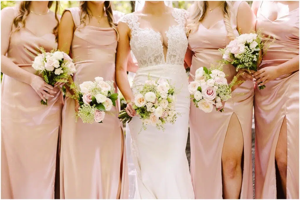 Snohomish Wedding Photographer GSquared Weddings A bride in a white gown stands with four bridesmaids in matching pink dresses, each holding a bouquet of white and pink flowers. The setting appears to be outdoors, with greenery in the background. Serving Seattle, Snohomish and Orlando