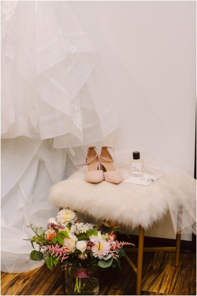 Snohomish Wedding Photographer GSquared Weddings A wedding dress hangs beside a small stool with pink heels, a bottle of perfume, and a note on it. Below, theres a bouquet of flowers in a glass vase on a wooden floor. Serving Seattle, Snohomish and Orlando