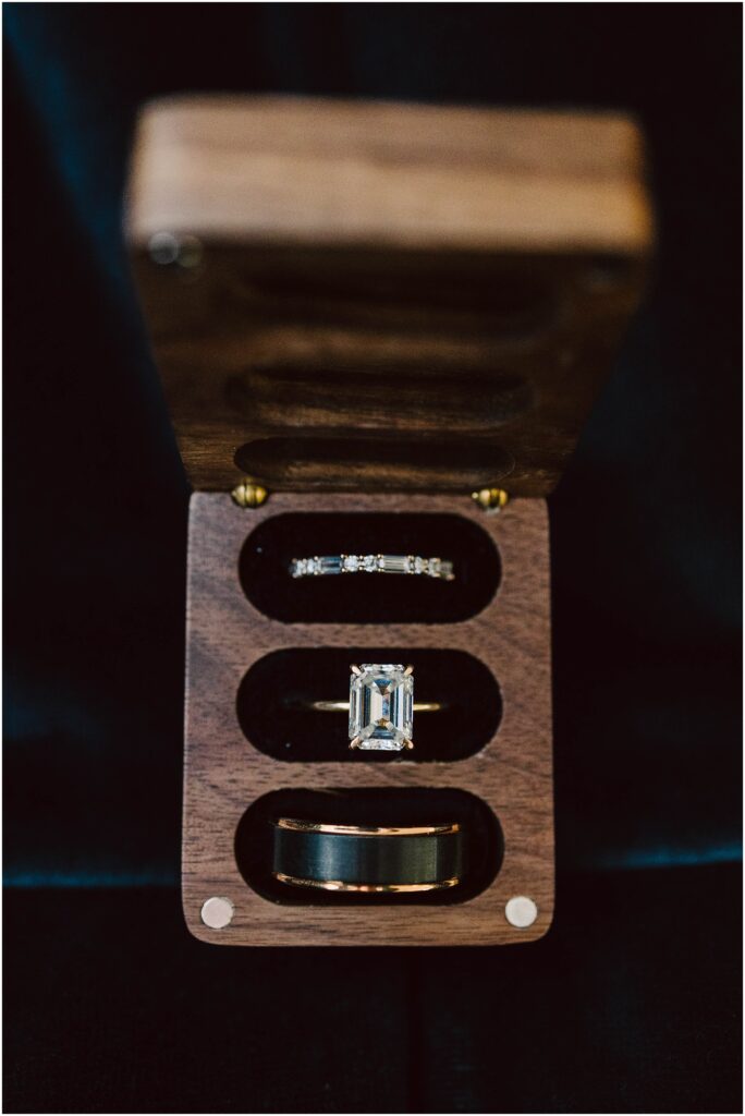Snohomish Wedding Photographer GSquared Weddings A wooden box with three compartments holds an emerald-cut diamond ring, a thin diamond-studded band, and a solid band, possibly a wedding ring set. The box is open, displaying the rings against a dark background. Serving Seattle, Snohomish and Orlando
