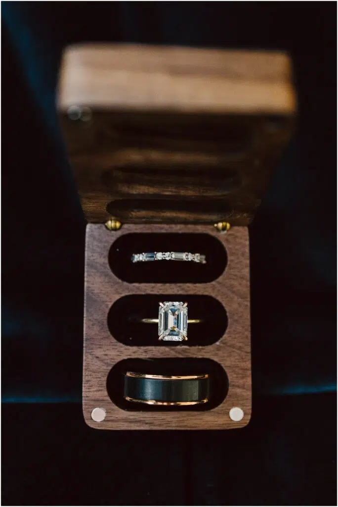 Snohomish Wedding Photographer GSquared Weddings A wooden box with three compartments holds an emerald-cut diamond ring, a thin diamond-studded band, and a solid band, possibly a wedding ring set. The box is open, displaying the rings against a dark background. Serving Seattle, Snohomish and Orlando