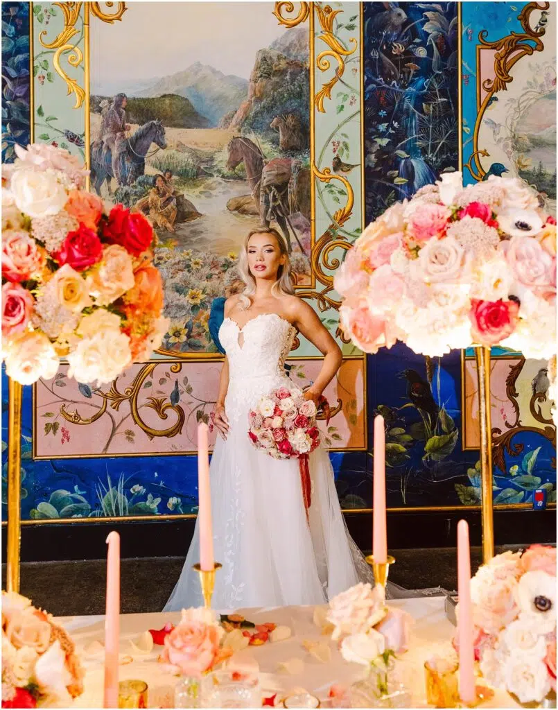 Snohomish Wedding Photographer GSquared Weddings A bride in a white gown holding a bouquet stands in front of a detailed mural depicting scenes and landscapes. The foreground features flower arrangements with pink and red roses and tall pink candles. Serving Seattle, Snohomish and Orlando