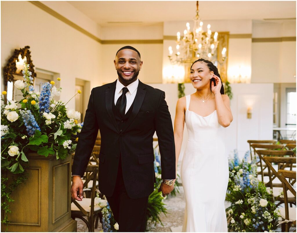 Snohomish Wedding Photographer GSquared Weddings A couple happily walks down the aisle after getting married. The groom wears a black suit and tie, while the bride is in a white dress. They are smiling, surrounded by floral arrangements and a chandelier in the background. Serving Seattle, Snohomish and Orlando