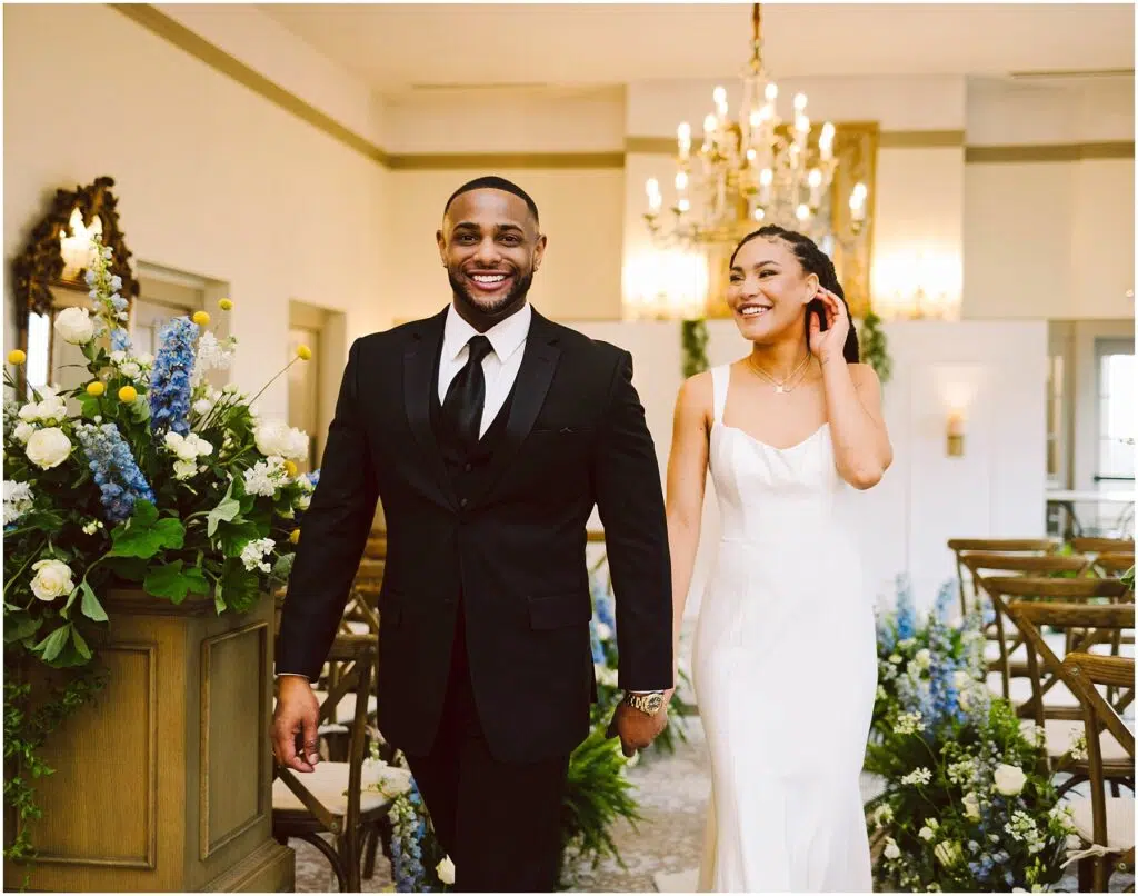 Snohomish Wedding Photographer GSquared Weddings A couple happily walks down the aisle after getting married. The groom wears a black suit and tie, while the bride is in a white dress. They are smiling, surrounded by floral arrangements and a chandelier in the background. Serving Seattle, Snohomish and Orlando