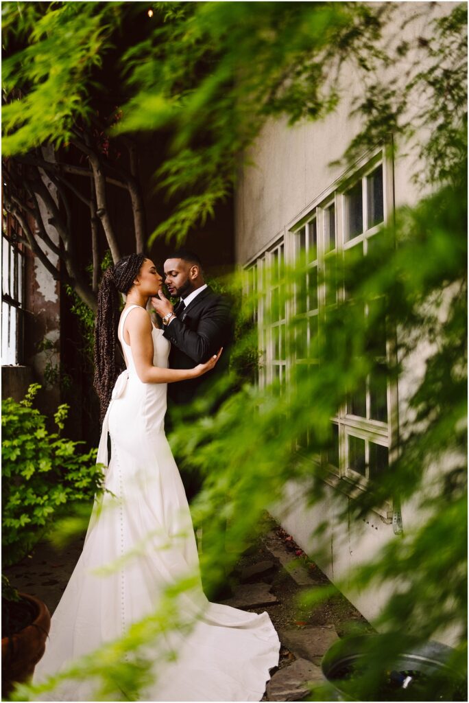Snohomish Wedding Photographer GSquared Weddings A bride in a white dress and a groom in a black suit share a close moment against a wall with windows. They are surrounded by lush greenery, creating an intimate and romantic atmosphere. Serving Seattle, Snohomish and Orlando