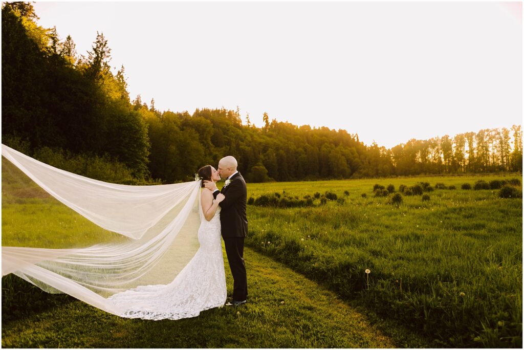 Snohomish Wedding Photographer GSquared Weddings A couple embraces in a sunlit meadow at sunset. The woman wears a flowing white gown with a long veil, while the man is in a dark suit. Trees and hills form the backdrop, bathed in a warm golden light. Serving Seattle, Snohomish and Orlando