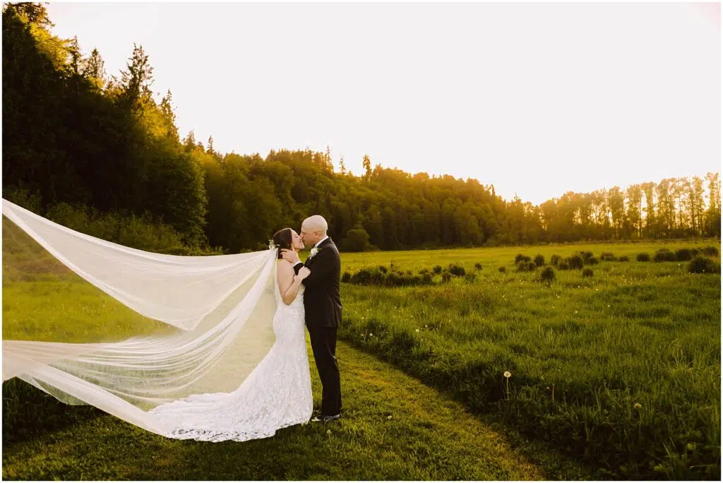 Snohomish Wedding Photographer GSquared Weddings A couple embraces in a sunlit meadow at sunset. The woman wears a flowing white gown with a long veil, while the man is in a dark suit. Trees and hills form the backdrop, bathed in a warm golden light. Serving Seattle, Snohomish and Orlando