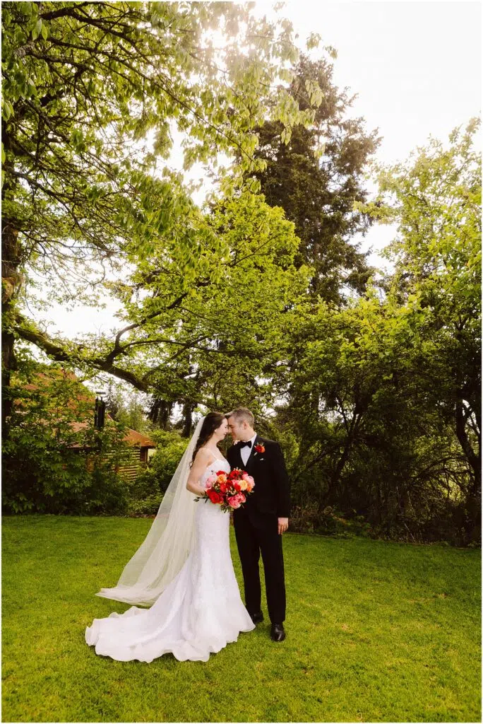 Snohomish Wedding Photographer GSquared Weddings A bride and groom share a kiss in a lush garden. The bride wears a white dress and veil, holding a bouquet of colorful flowers. The groom is in a black suit. They stand on vibrant green grass surrounded by trees under a bright sky. Serving Seattle, Snohomish and Orlando