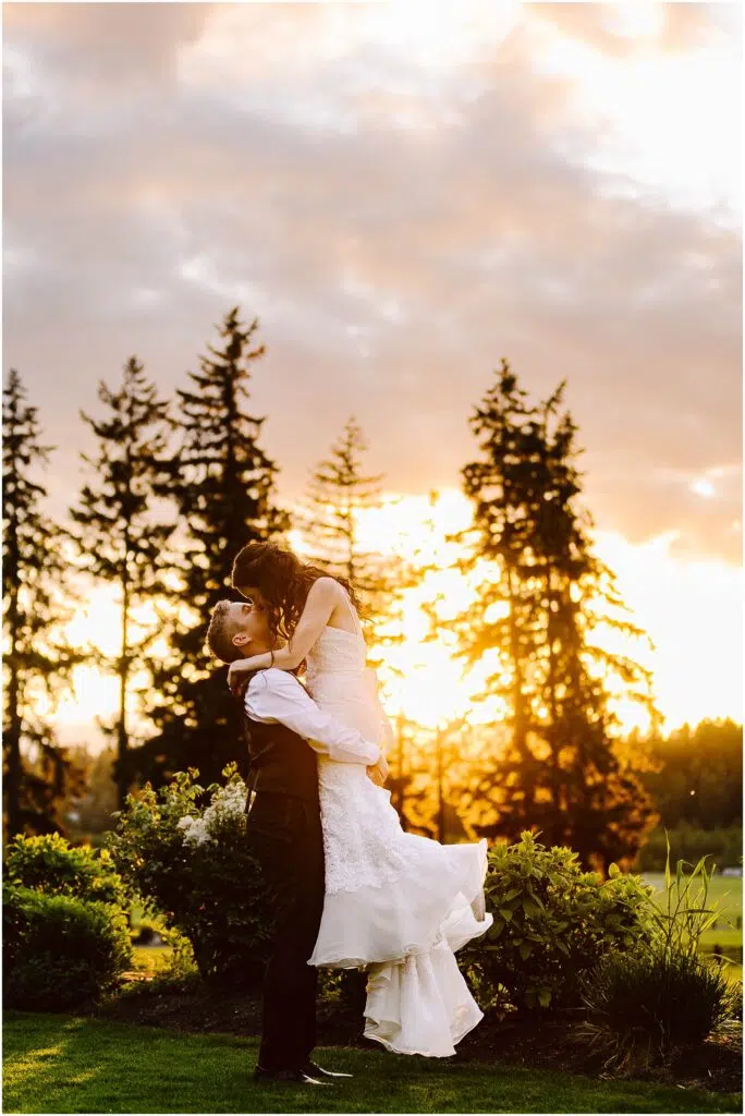 Snohomish Wedding Photographer GSquared Weddings A couple is embracing outdoors at sunset, with the man lifting the woman. They are surrounded by greenery, and tall trees are silhouetted against the vibrant sky. The woman is wearing a white dress, and the man is in a vest and trousers. Serving Seattle, Snohomish and Orlando