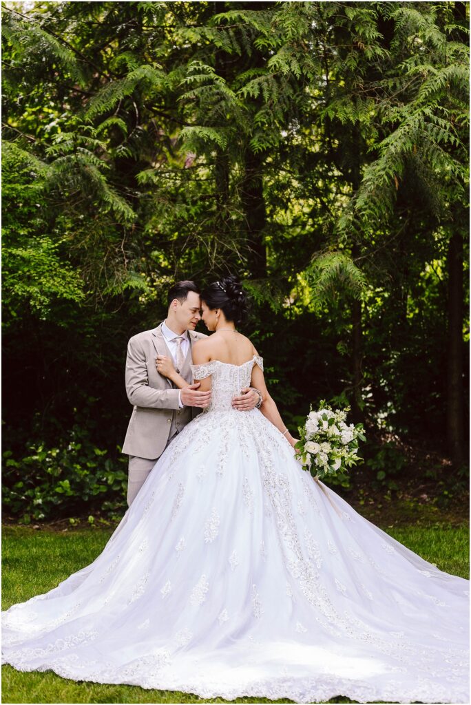 Snohomish Wedding Photographer GSquared Weddings A couple stands in a lush garden, embracing under tall trees. The bride wears an elegant white gown, holding a bouquet of flowers. The groom is in a light-colored suit. They share a tender moment, facing each other closely. Serving Seattle, Snohomish and Orlando