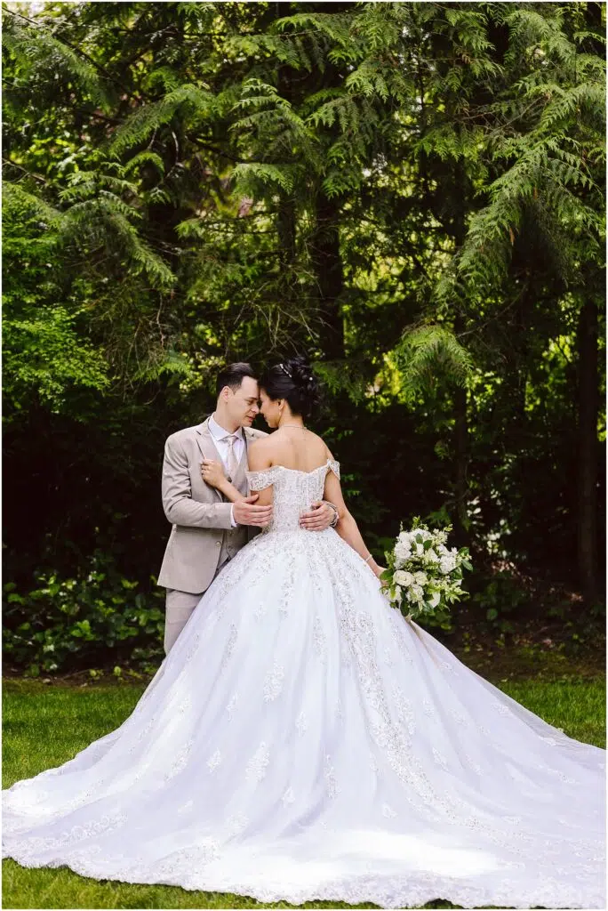 Snohomish Wedding Photographer GSquared Weddings A couple stands in a lush garden, embracing under tall trees. The bride wears an elegant white gown, holding a bouquet of flowers. The groom is in a light-colored suit. They share a tender moment, facing each other closely. Serving Seattle, Snohomish and Orlando