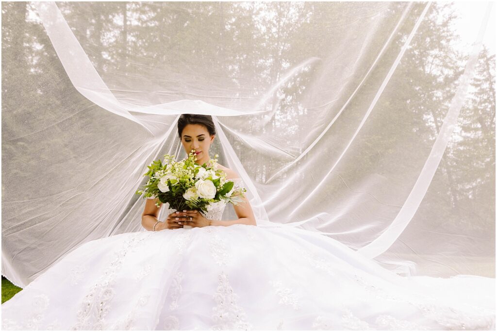 Snohomish Wedding Photographer GSquared Weddings A bride in an elegant white gown sits holding a bouquet of white and green flowers. Her veil is spread out behind her, creating a delicate backdrop. The scene is set outdoors with a blurred background of trees. Serving Seattle, Snohomish and Orlando
