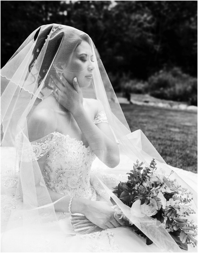 Snohomish Wedding Photographer GSquared Weddings Bride in an intricately beaded gown sits outdoors, partially covered by a sheer veil. She holds a bouquet and rests one hand on her neck, looking serene. The background features a blurred natural setting. Black and white image. Serving Seattle, Snohomish and Orlando