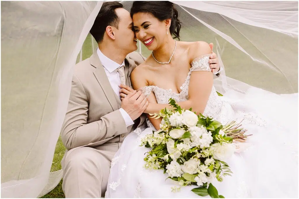 Snohomish Wedding Photographer GSquared Weddings Bride and groom sitting under a veil, smiling. The bride is in an off-shoulder white dress holding a bouquet of white flowers. The groom is in a beige suit. Theyre on grass, sharing an intimate, joyful moment. Serving Seattle, Snohomish and Orlando