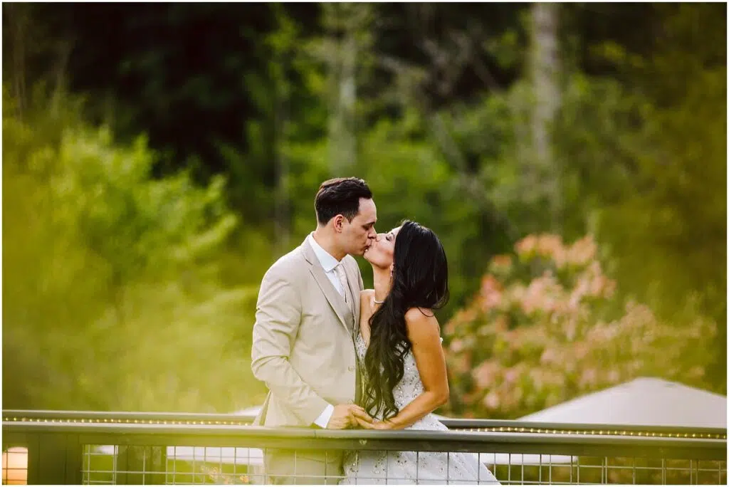Snohomish Wedding Photographer GSquared Weddings A couple kisses on a bridge in a lush, green park. The man, in a beige suit, and the woman, in a white dress, share an intimate moment surrounded by greenery and blurred floral backgrounds. Serving Seattle, Snohomish and Orlando