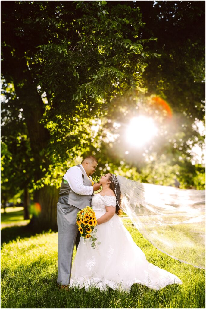 Snohomish Wedding Photographer GSquared Weddings A bride and groom stand under a large tree with sunlight streaming through the leaves. The bride holds a bouquet of sunflowers and her veil flows in the breeze. Both are gazing at each other, smiling warmly. Serving Seattle, Snohomish and Orlando