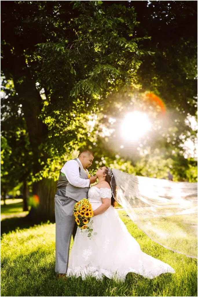 Snohomish Wedding Photographer GSquared Weddings A bride and groom stand under a large tree with sunlight streaming through the leaves. The bride holds a bouquet of sunflowers and her veil flows in the breeze. Both are gazing at each other, smiling warmly. Serving Seattle, Snohomish and Orlando