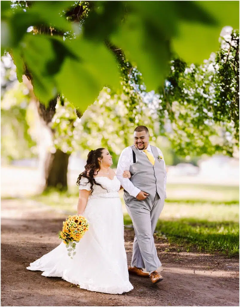 Snohomish Wedding Photographer GSquared Weddings A bride in a white dress holds a sunflower bouquet, walking hand in hand with a groom in a gray suit and suspenders. They are smiling at each other under lush green trees, sharing a joyful moment on a sunlit path. Serving Seattle, Snohomish and Orlando