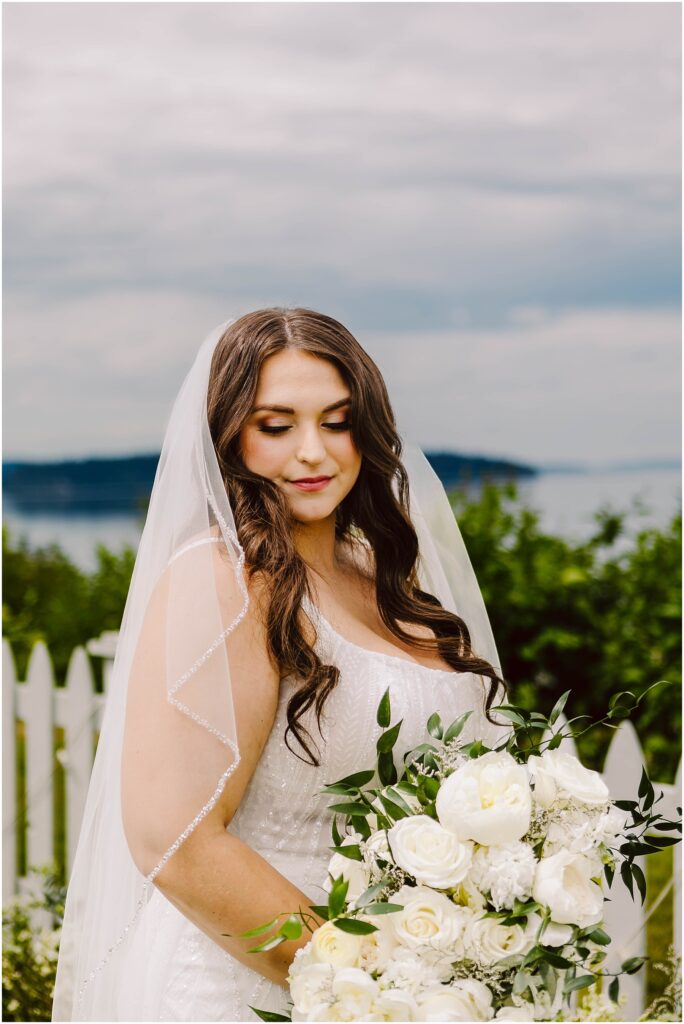 Snohomish Wedding Photographer GSquared Weddings A bride stands outdoors with a white picket fence and waterfront view in the background. She holds a bouquet of white flowers and greenery, wearing a white dress and veil. The sky is overcast. Serving Seattle, Snohomish and Orlando