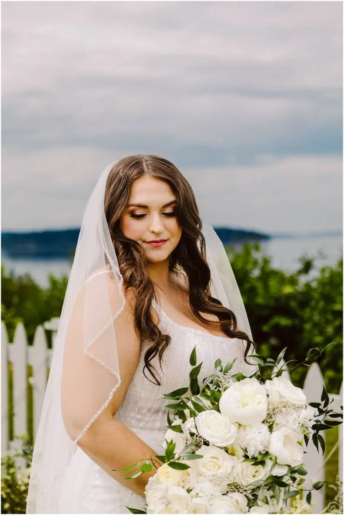 Snohomish Wedding Photographer GSquared Weddings A bride stands outdoors with a white picket fence and waterfront view in the background. She holds a bouquet of white flowers and greenery, wearing a white dress and veil. The sky is overcast. Serving Seattle, Snohomish and Orlando
