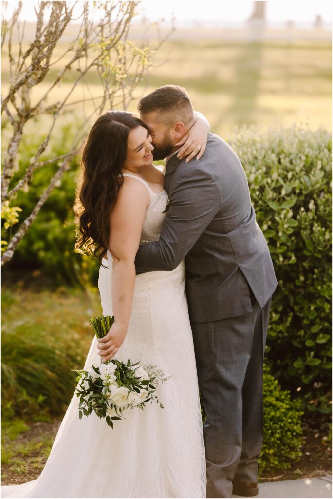 Snohomish Wedding Photographer GSquared Weddings A couple embraces outdoors in a garden setting. The bride is wearing a white dress and holding a bouquet of white flowers. The groom is in a gray suit. They are smiling and appear joyful, with the sun softly illuminating the scene. Serving Seattle, Snohomish and Orlando