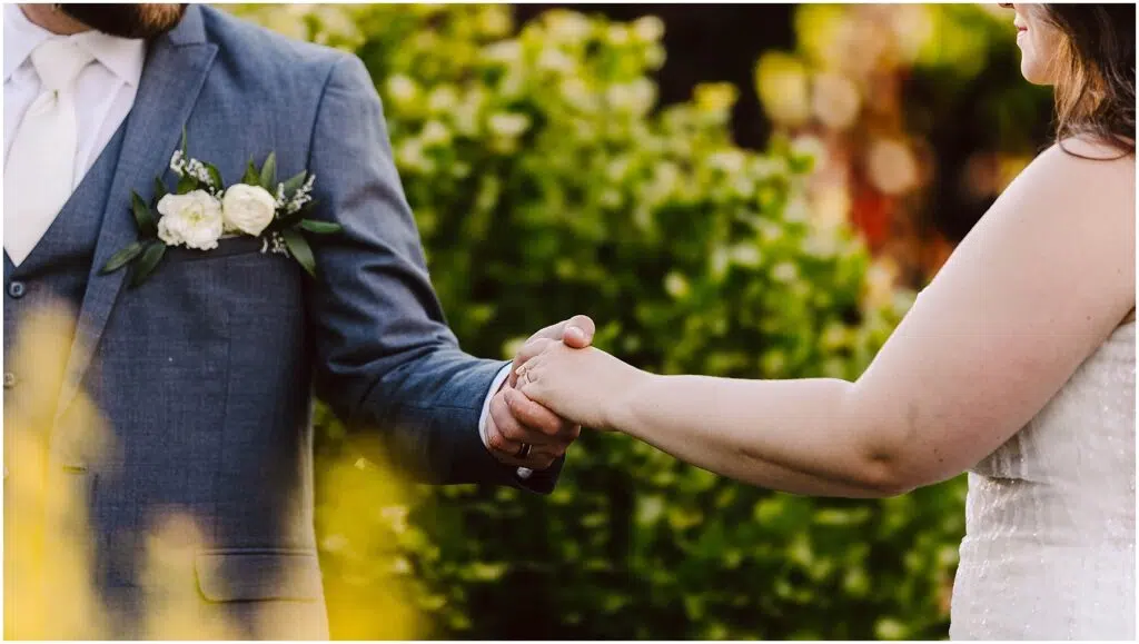 Snohomish Wedding Photographer GSquared Weddings A couple holding hands, dressed formally. The person on the left wears a blue suit with a white boutonniere. The person on the right is in a white dress. The background is filled with greenery. Serving Seattle, Snohomish and Orlando