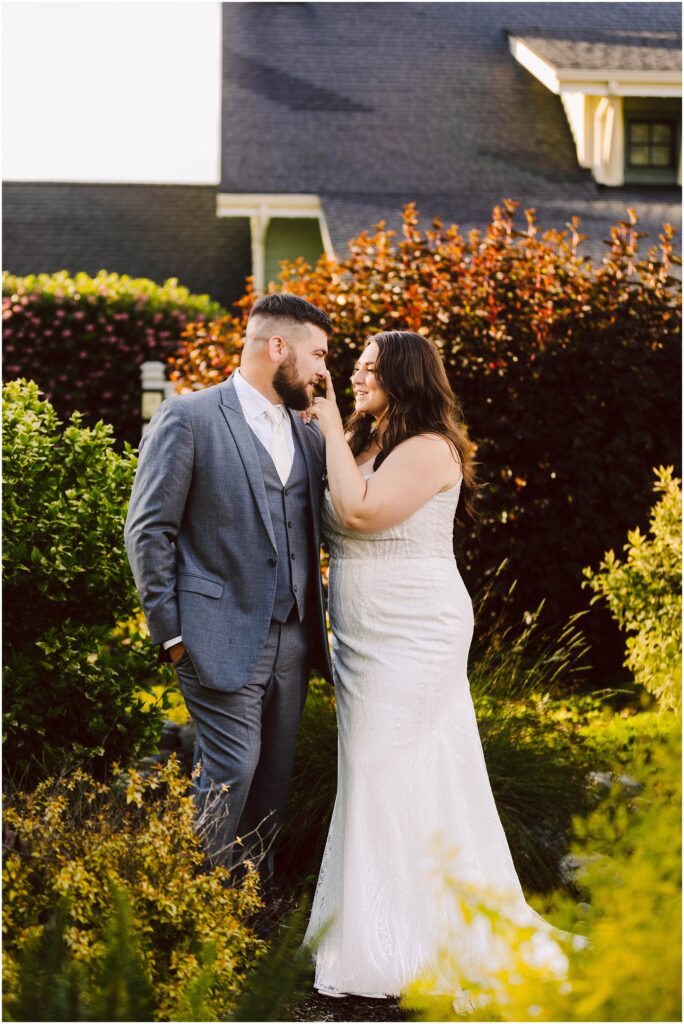 Snohomish Wedding Photographer GSquared Weddings A couple stands in a garden. The man is wearing a gray suit and the woman is in a white wedding dress. She is touching his beard, and they are smiling at each other. The background has bushes and a building with a steeply pitched roof. Serving Seattle, Snohomish and Orlando