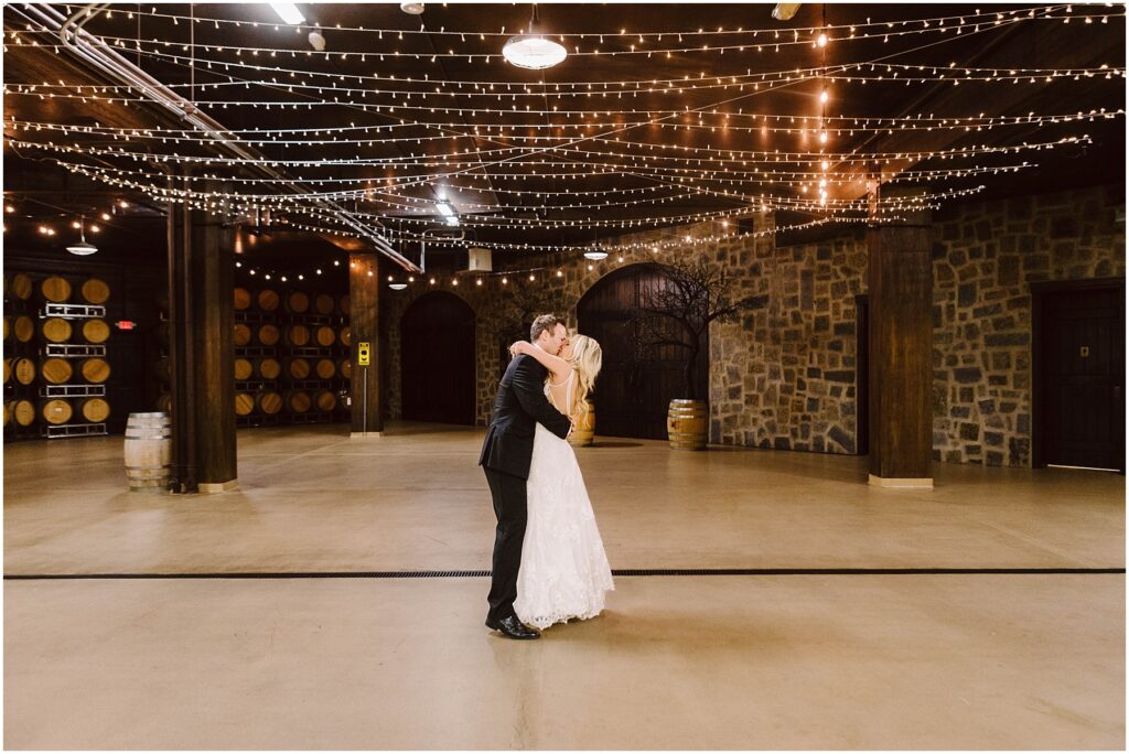 Snohomish Wedding Photographer GSquared Weddings A bride and groom share a kiss in an empty, warmly lit winery cellar adorned with string lights. Wine barrels line one wall, and the stone walls create a rustic ambiance. The couple stands in the center of the spacious, festive area. Serving Seattle, Snohomish and Orlando