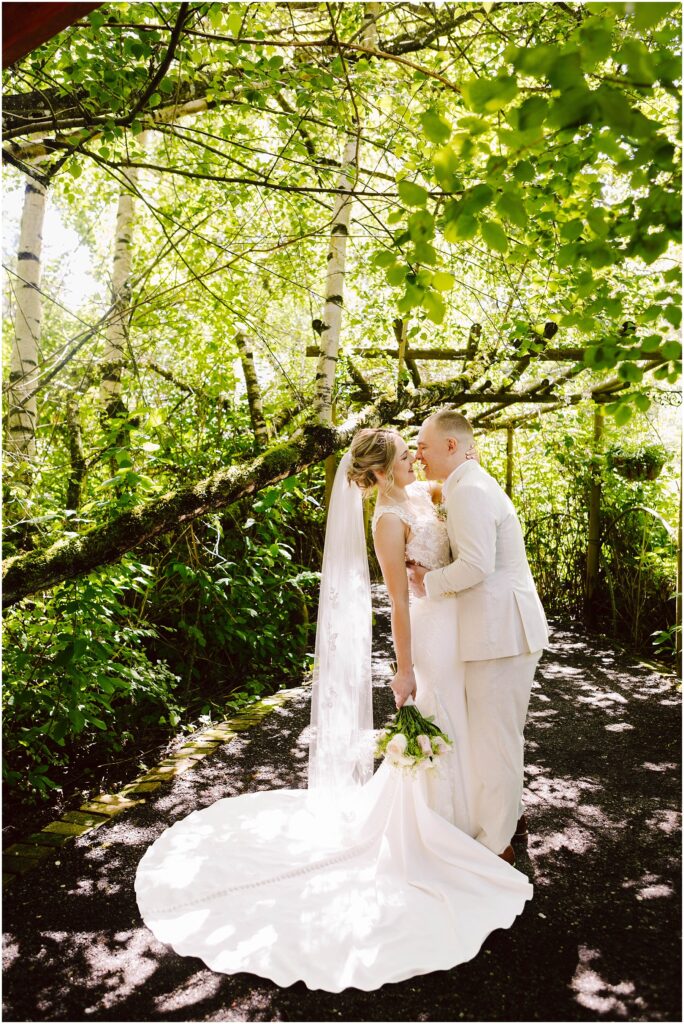 Snohomish Wedding Photographer GSquared Weddings A couple in wedding attire shares a kiss under sun-dappled trees. The bride holds a bouquet and wears a long veil, while the groom is in a light suit. Sunlight filters through the lush green leaves, creating a serene and romantic scene. Serving Seattle, Snohomish and Orlando