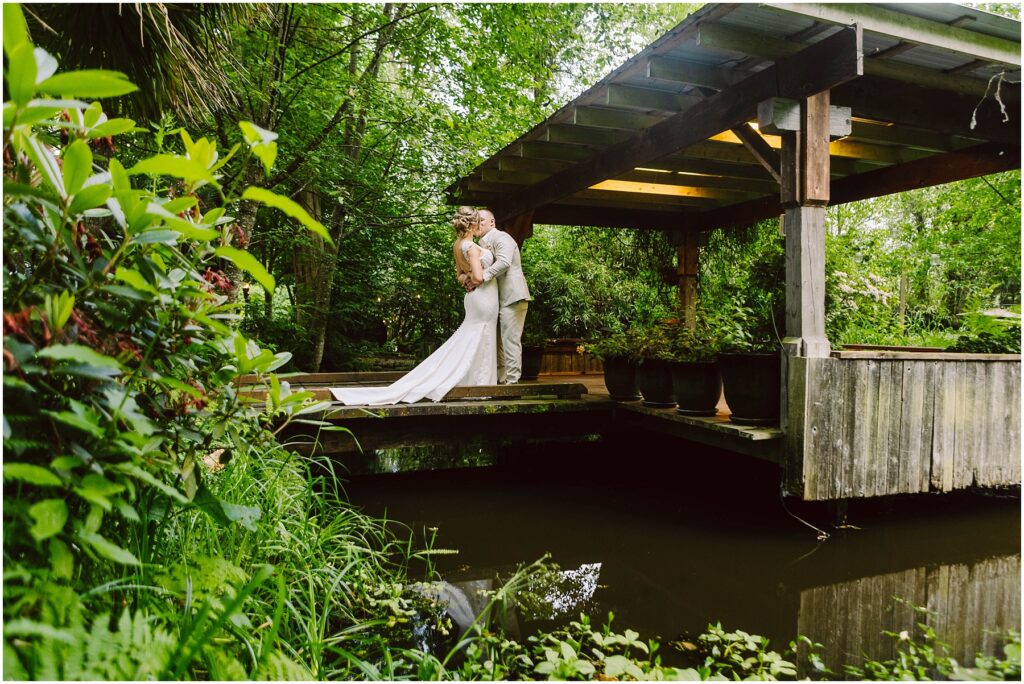 Snohomish Wedding Photographer GSquared Weddings A couple in wedding attire embraces under a wooden gazebo surrounded by lush greenery. A small pond with reflective water is in the foreground, adding to the serene and romantic setting. Serving Seattle, Snohomish and Orlando
