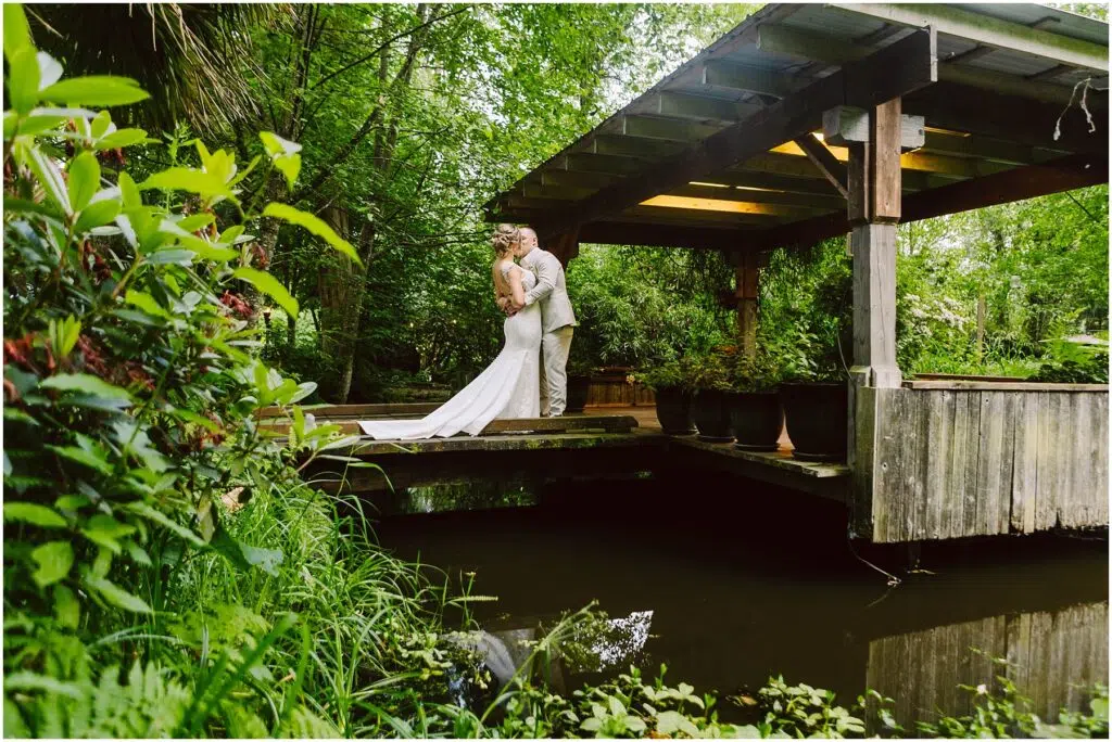 Snohomish Wedding Photographer GSquared Weddings A couple in wedding attire embraces under a wooden gazebo surrounded by lush greenery. A small pond with reflective water is in the foreground, adding to the serene and romantic setting. Serving Seattle, Snohomish and Orlando