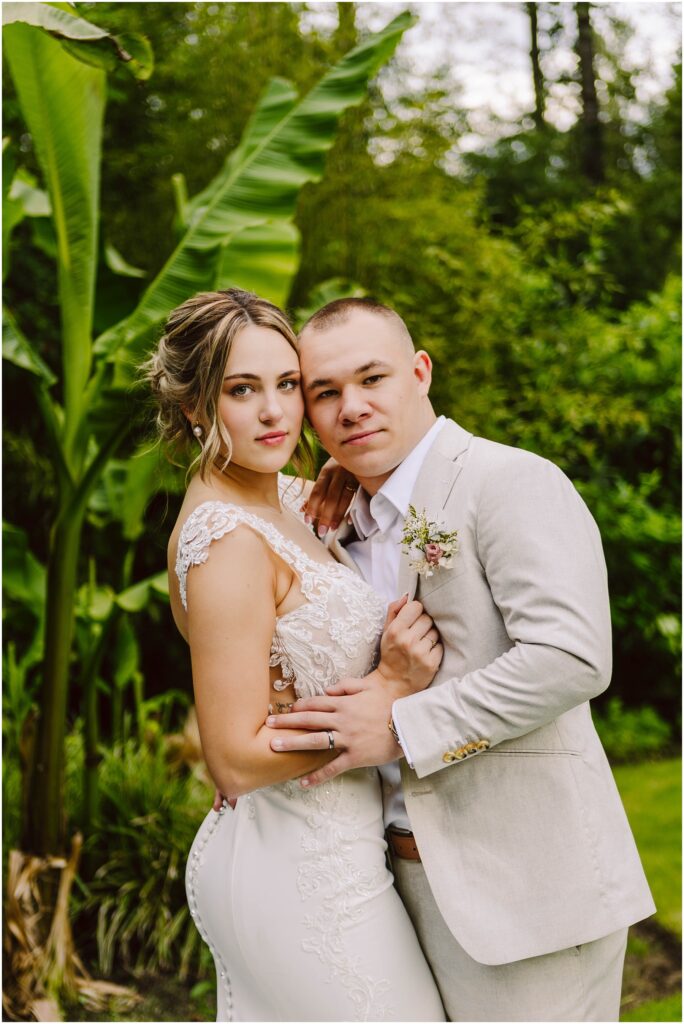 Snohomish Wedding Photographer GSquared Weddings A bride in a white lace dress and a groom in a light gray suit pose together in a lush garden setting. They are embracing, looking at the camera, with large green leaves visible in the background. Serving Seattle, Snohomish and Orlando