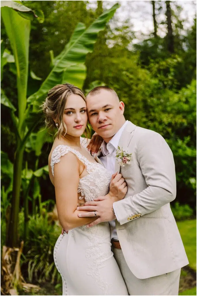 Snohomish Wedding Photographer GSquared Weddings A bride in a white lace dress and a groom in a light gray suit pose together in a lush garden setting. They are embracing, looking at the camera, with large green leaves visible in the background. Serving Seattle, Snohomish and Orlando
