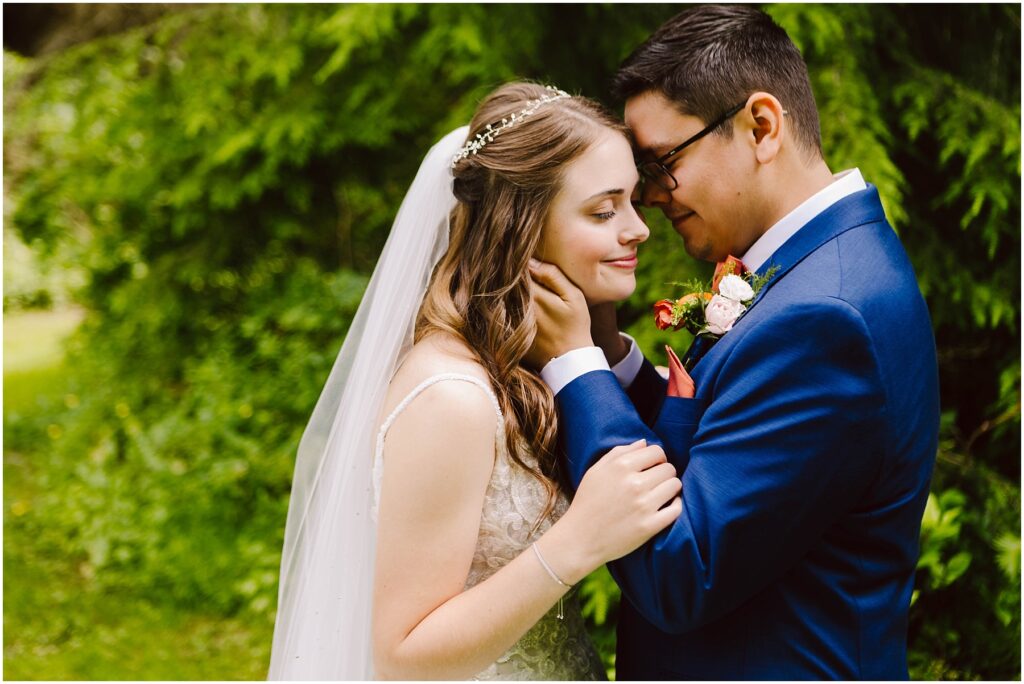 Snohomish Wedding Photographer GSquared Weddings A bride in a white gown and veil and a groom in a blue suit share a tender moment outdoors. The groom gently touches the brides face as they both smile with their eyes closed, surrounded by lush greenery. Serving Seattle, Snohomish and Orlando