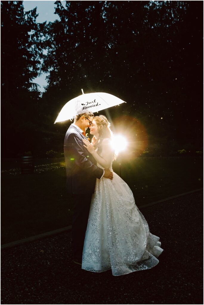 Snohomish Wedding Photographer GSquared Weddings A bride and groom embrace under a glowing umbrella that reads Just Married, with a soft light shining behind them. They are standing on a dark path surrounded by trees. The bride is wearing a white gown, and the groom is in a suit. Serving Seattle, Snohomish and Orlando
