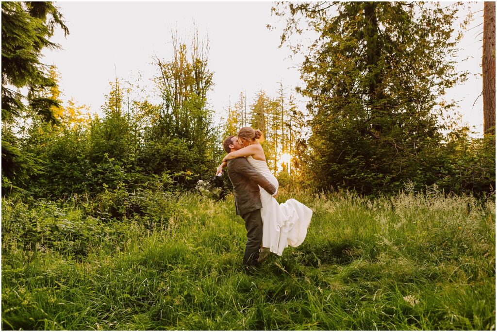 Snohomish Wedding Photographer GSquared Weddings A couple in wedding attire embrace joyfully in a lush, green forest clearing. The sun sets in the background, casting a warm glow over the scene. The bride is lifted off the ground by the groom, surrounded by tall trees and grass. Serving Seattle, Snohomish and Orlando